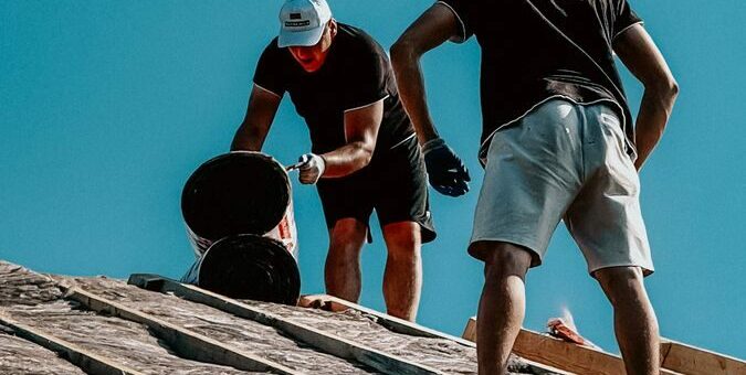 Two construction workers apply roofing materials under a bright blue sky, showcasing teamwork and labor.