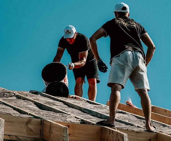 Two construction workers apply roofing materials under a bright blue sky, showcasing teamwork and labor.